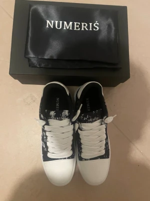 Svarta och vita sneakers från Numeris - Snygga sneakers från Numeris i svart och vitt med coolt mönster på sidorna. Skorna har vita snören, vit tå och häl samt svart insida. Ovandelen verkar vara i textil och syntet, sulan är vit och platt. Perfekta för en trendig streetstyle-look.