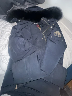 Svart dunjacka med pälskrage Moose Knuckles - Snygg svart dunjacka från Moose Knuckles med stor fluffig pälskrage och dragkedja framtill. Jackan har logga på ärmen och flera detaljer i metall. Perfekt för kalla vinterdagar och riktigt varm.