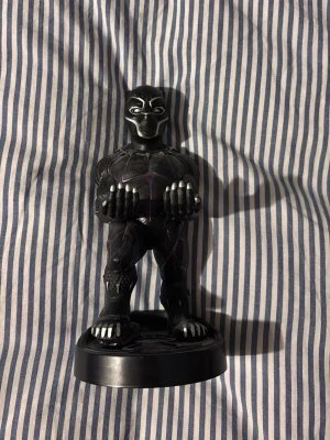Black Panther figur med hållare - Cool Black Panther figur i svart plast med lila detaljer och silverfärgade klor. Figuren står på en rund svart bas och har armarna framåt som en hållare, perfekt för att ställa mobil eller handkontroll på. Perfekt för Marvel-fans!