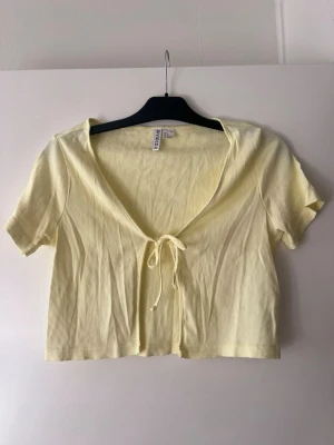 Ljusgul ribbad croppad topp från Divided - Superfin ljusgul croppad topp från Divided med korta ärmar och v-ringning. Toppen är ribbad och har en söt knytning framtill. Perfekt för dig som gillar en enkel men trendig stil.
