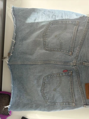 Ljusblå jeanskjol från Levi's - Trendig ljusblå jeanskjol från Levi's med råa kanter och slitna detaljer. Klassisk femficksmodell med knapp- och dragkedjestängning framtill. Perfekt för dig som gillar en avslappnad och cool stil.