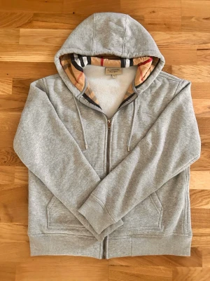 Grå zip-hoodie från Burberry - Snygg grå hoodie från Burberry med dragkedja och klassiskt rutigt foder i huvan. Mjuk bomullsblandning, två fickor fram och justerbar dragsko i huvan. Perfekt för en chill och stilren look.