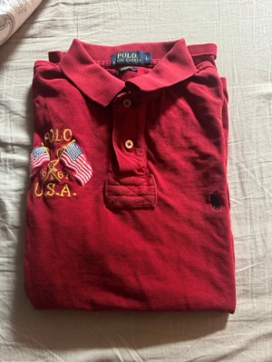 Röd pikétröja från Polo Ralph Lauren - Röd pikétröja från Polo Ralph Lauren med broderad logga på bröstet och USA-detaljer med flaggor. Klassisk krage och knappar framtill. Har ett litet hål vid vänstra armhålan 