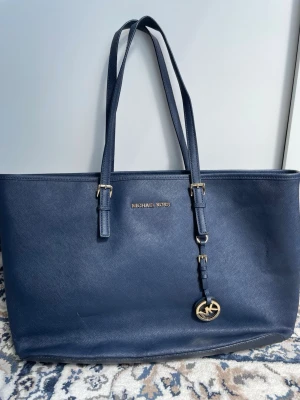 Mörkblå shoppingväska Michael Kors - Snygg mörkblå shoppingväska från Michael Kors i slätt läder med guldfärgade detaljer och logotyp framtill. Väskan har dubbla axelremmar och en klassisk, rymlig design. Perfekt för dig som vill ha en stilren och praktisk väska med lyxig känsla.