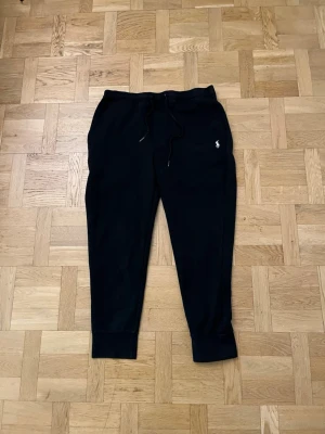 Svarta mjukisbyxor från Polo Ralph Lauren - Svarta mjukisbyxor från Polo Ralph Lauren med dragsko i midjan, resår vid benslut och klassisk vit logga broderad på vänster sida. Byxorna har fickor fram och en bakficka. Perfekta för en avslappnad och stilren look.