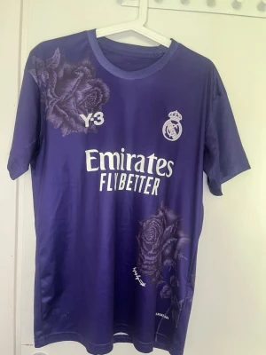 Lila Real Madrid fotbollströja Y-3 - Snygg lila matchtröja från Real Madrid i samarbete med Y-3. Tröjan har tryckta rosor, klubbmärke och sponsorlogga på bröstet. Tillverkad i lätt och ventilerande material, perfekt för fotboll eller träning. Rund hals och korta ärmar ger en sportig look. Kom privat vid funderingar 