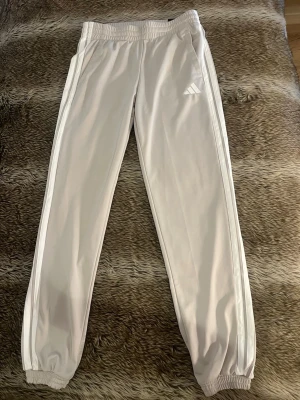 Vita träningsbyxor från Adidas - Säljer ett par vita träningsbyxor från Adidas med klassiska tre ränder längs benen och elastisk midja. Byxorna har resår vid bensluten och är tillverkade i ett lätt, sportigt material som passar perfekt till träning eller chill. Adidas-logga på vänster lår.