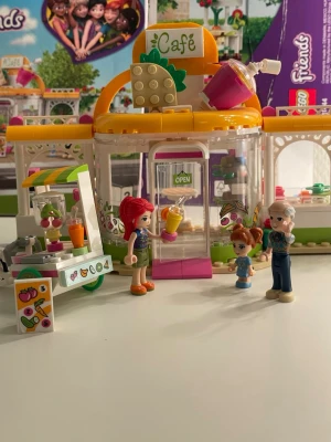 LEGO Friends Juice Café set - Färgglatt LEGO Friends Juice Café med tillhörande juicevagn, uteservering och tre minifigurer och om du vill ha djur som ekorre eller hund så ingår de. Setet har detaljer som frukt, drycker och skyltar. Byggsatsen är i plast och har inslag av gult, vitt, grönt och rosa. Perfekt för dig som gillar att bygga och leka café. Men hör av er om ni vill se mer av mina samlingar.