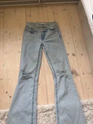 Ljusblå bootcut jeans med slitningar - Säljer ett par ljusblå bootcut jeans med slitna detaljer på båda knäna och snygga röda broderier på bakfickorna. Jeansen har klassisk femficksdesign och är tillverkade i denim. Perfekta för en avslappnad och trendig look.