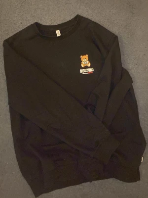 Svart Moschino sweatshirt med björn - Cool svart sweatshirt från Moschino med deras ikoniska björntryck och logga på bröstet. Tröjan har rund halsringning och långa ärmar. Perfekt för dig som gillar streetwear och vill sticka ut med en snygg detalj.