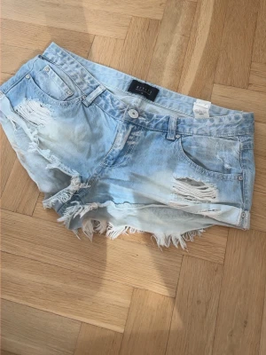 Ljusblå denimshorts med fransar - Säljer ett par ljusblå denimshorts från Bik Bok med slitna detaljer och fransig kant. Shortsen har klassisk femficksmodell, låg midja och en avslappnad passform. Perfekta för varma dagar och en chill look.