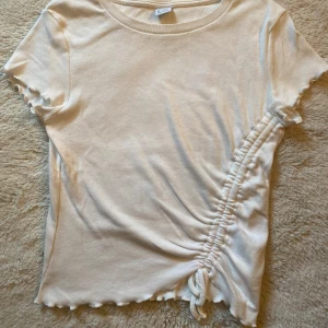 Vit ribbad t-shirt med snörning - Superfin vit ribbad t-shirt från Lindex med korta ärmar och snörning på sidan som ger en snygg rynkad effekt. Modell med vågiga kanter på ärmar och nederkant. 