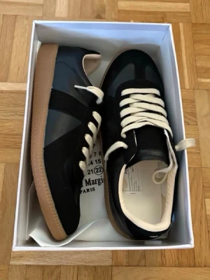 Maison Margiela gats storlek 43 - Svarta sneakers från Maison Margiela med beige sula och vita skosnören. Skorna har en klassisk låg siluett, ovandel i läder och mocka samt diskreta detaljer. Perfekta för en stilren och trendig look.