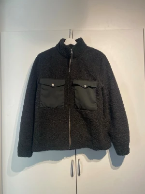 These Glory Days Teddyjacka - Mysig mörkgrön teddyjacka med dragkedja framtill och två stora fickor med lock och knapp på bröstet från These Glory Days. Jackan har hög krage och är gjord i fluffigt teddy-material med detaljer i nylon på fickorna. Storlek M, TTS Small. Väldigt bra skick 