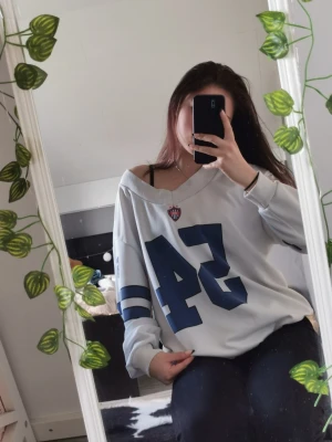 Vit oversized tröja med blå tryck - Vit oversized tröja med blå siffror och ränder på ärmarna. Tröjan har en djup v-ringning och sportig collegekänsla, perfekt för en avslappnad stil. Stora siffror framtill och emblem vid halsen ger en cool look.