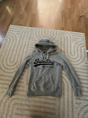 Grå Superdry hoodie med tryck - Snygg grå hoodie från Superdry med svart logotyp och japanska tecken på bröstet. Tröjan har huva med vita snören och en stor magficka. Perfekt för en avslappnad och cool stil.
