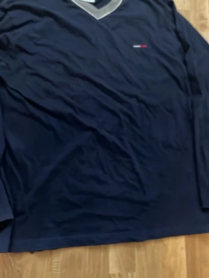 Mörkblå v-ringad tröja Tommy Hilfiger - Mörkblå långärmad tröja från Tommy Hilfiger med v-ringning och grå kant runt halsen. Liten broderad logga på bröstet. Perfekt för en clean och stilren look.