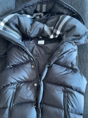 Burberry väst - Snygg svart dunväst från burberry,  och använda en gång. Insidan av huvan har ett grått och vitt rutigt foder. Västen har två dragkedjefickor framtill och stängs med både dragkedja och tryckknappar och kan göras till en jacka. Perfekt för sommaren med en skön och snygg väst