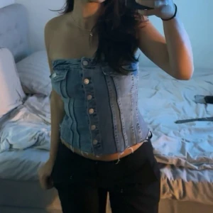 Blå denim korsett-topp med knappar - Säljer en snygg blå korsett-topp i denim med knappar framtill och dekorativa bröstfickor. Toppen är axelbandslös och har en croppad passform. Skit snygg med ett par denim jeans 😍 den har en liten rosa fläck intill ( läppstift) men som går att tvätta bort och inget som syns utifrån