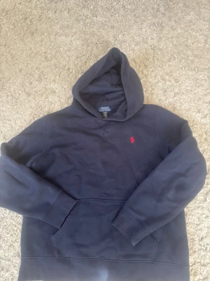 Mörkblå hoodie från Polo Ralph Lauren - Snygg mörkblå hoodie från Polo Ralph Lauren med klassisk röd logga broderad på bröstet. Hoodien har huva och en stor magficka framtill. Tillverkad i mjukt material som känns skönt mot huden. Perfekt för en avslappnad och stilren look. Största barnstorlek som sitter som xs/S