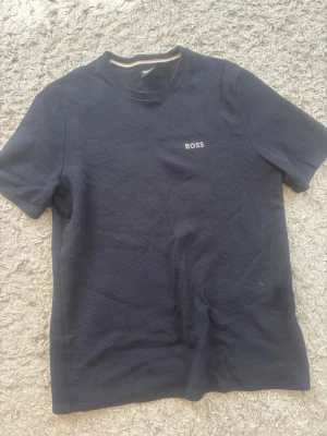 Svart t-shirt från BOSS - Snygg svart t-shirt från BOSS med diskret logga på bröstet. T-shirten har rund halsringning och korta ärmar. Materialet känns mjukt och har en lätt textur som ger en cool känsla till outfiten. 