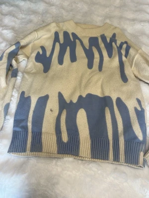 Beige stickad tröja med blått mönster - Cool beige stickad tröja med unikt blått drip-mönster. Tröjan har rund halsringning och ribbade muddar vid ärmslut och nederkant. Perfekt för dig som vill sticka ut med en trendig och konstnärlig look.