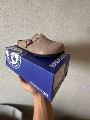 Beige Birkenstock Boston tofflor - Säljer ett par beige Birkenstock Boston tofflor i mocka med justerbart spänne och klassisk korksula. Yttersulan är i svart gummi med mönster för bra grepp. Perfekta för dig som gillar stilrena och bekväma skor.