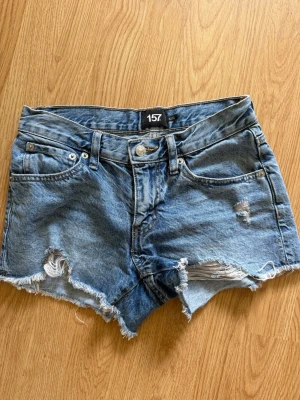 Blå denimshorts med slitningar - Säljer ett par blå denimshorts från 157 med råa kanter och slitna detaljer. Klassisk femficksmodell med knapp och dragkedja framtill. Perfekta för varma dagar och ger en avslappnad look. Säljer dom på grund av att de är för små, har därför ingen bild med dom på heller.