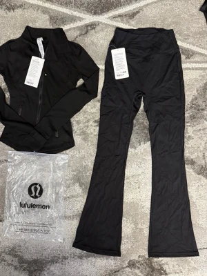 Svart träningsset från Lululemon - Snyggt svart träningsset från Lululemon med en åtsittande Define Jacket med dragkedja och hög krage samt ett par utsvängda träningsbyxor. Materialet är stretchigt och följsamt, perfekt för gymmet eller yoga. Diskret logga och stilren design.