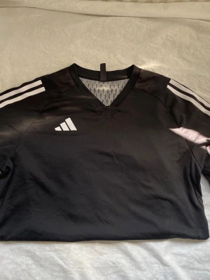 Svart Adidas sporttröja med ränder - Säljer en svart sporttröja från Adidas med klassiska vita ränder på axlarna och vitt Adidas-logga på bröstet. Tröjan har V-ringning med meshdetalj och är tillverkad i ett lätt, snabbtorkande material. Perfekt för träning eller match!