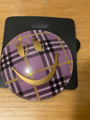 Hårsnoddar med rutigt smiley-motiv - Två hårsnoddar med stora runda dekorationer i rutigt mönster, en i lila och en i rosa. Båda har ett guldigt smiley-ansikte med ögonfransar. Perfekt för att ge en lekfull touch till din frisyr. 25kr/st , helt ny 