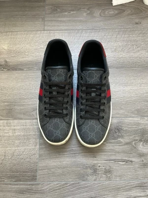 Gucci sneakers med monogram och röd rand - Snygga Gucci sneakers i svart med klassiskt GG-monogram över hela skon och en röd/blå rand på sidan. Skorna har svarta skosnören, vit sula och är tillverkade i canvas med läderdetaljer. Perfekta för dig som vill ha en stilren och exklusiv look.