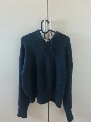 Mörkblå stickad hoodie med huva - Mörkblå stickad hoodie med huva och dragsko. Man ser att tröjan är använd för att den är lite nopprig. Materialet är lite stickigt, men man vänjer sig efter ett tag.