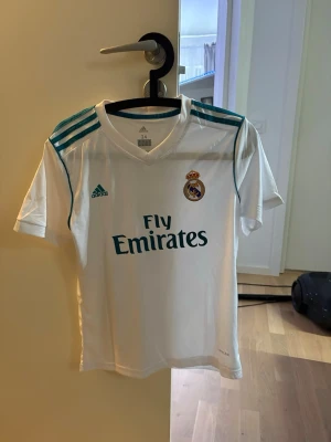 Real Madrid Ronaldo matchtröja - Säljer en vit Real Madrid matchtröja från Adidas med turkosa detaljer och klubbmärke på bröstet. Tryck med 'Fly Emirates' på framsidan och 'Ronaldo 7' på ryggen. Klassisk V-ringning och korta ärmar, perfekt för fotbollsträning eller att bära till vardags.