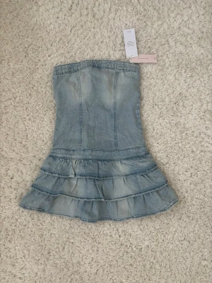 Ljusblå axelbandslös jeansklänning - Trendig axelbandslös kort jeansklänning i ljusblå denim med volangdetaljer nertill och dragkedja bak. Perfekt för dig som vill ha en cool och somrig look med en feminin touch.