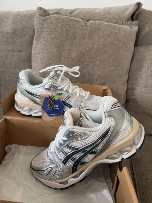 ASICS vita och silver sneakers - Snygga ASICS sneakers i vitt och silver med svarta detaljer. Skorna har en sportig design med mesh och syntetmaterial, samt en chunky sula för extra komfort. Perfekta för dig som gillar streetstyle och sportig look. Klassisk ASICS-logga på sidan och vita skosnören. det är storlek 43 men små i storleken så har du 42 så passar det .