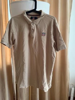 Beige pikétröja från Vinson Polo Club - Snygg beige pikétröja från Vinson Polo Club med klassisk krage och knappar framtill. Tröjan har korta ärmar och ett broderat märke på bröstet med röd, vit och svart detalj. Perfekt till en avslappnad stil.
