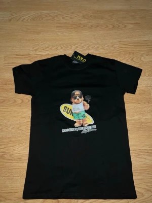 Svart t-shirt Polo Ralph Lauren björn - Svart t-shirt från Polo Ralph Lauren med ikonisk björn-tryck på bröstet. Björnen har solglasögon, keps och håller en surfbräda. T-shirten har rund hals och korta ärmar. Perfekt för en avslappnad och cool stil.