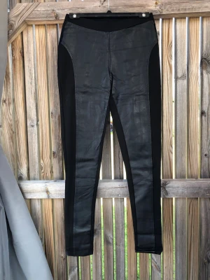 Svarta leggings med fuskskinnpaneler - Snygga svarta leggings med fuskskinnpaneler framtill och stretchigt tyg på sidor och baksida. Hög midja och smal passform som ger en cool och trendig look. Perfekta till oversized tröjor eller crop tops. Materialmix av fuskskinn och stretchtyg.