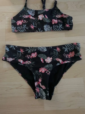 Blommig bikini från KappAhl - Snygg bikini från KappAhl med svart botten och tropiskt blommönster i rosa och grönt. Överdelen har smala axelband och underdelen är klassisk i modellen. Perfekt för strandhäng eller poolen!