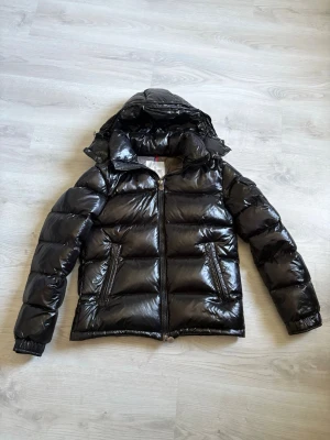 Svart glansig dunjacka med huva - Säljer en svart, glansig dunjacka med avtagbar huva och två dragkedjeförsedda fickor framtill. Jackan har breda, quiltade paneler och dragkedja hela vägen. Insidan är grå och har en serietidningslapp. Perfekt för kalla vinterdagar.