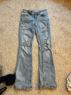 Lågmidjande jeans med hål - Säljer ett par ljusblå jeans med bootcut-modell och flera slitningar och hål på benen för en trendig look. Jag har gjort hålen själv🩷🩷