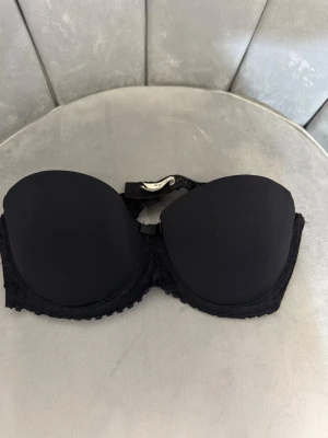 Svart axelbandslös bh Préambule 80C - Snygg svart axelbandslös bh från Préambule i storlek 80C. Bh:n har vadderade kupor och spetsdetaljer längs kanterna för en elegant look. Perfekt under toppar och klänningar med bara axlar.