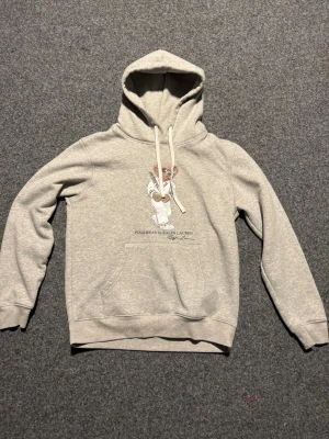 Grå hoodie med Polo Bear print - Snygg grå hoodie från Ralph Lauren med Polo Bear-motiv på bröstet. Klassisk känguruficka framtill, ribbade muddar och justerbar huva med vita snören. Perfekt för en chill och avslappnad stil. Mjuk bomullsblandning som känns skön mot huden.