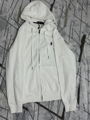 Vit hoodie från Polo Ralph Lauren - Vit hoodie med dragkedja från Polo Ralph Lauren. Klassisk design med huva och dragsko, två fickor framtill och broderad logga på bröstet. Perfekt för en avslappnad och stilren look. Har bara använt den 2 gånger.