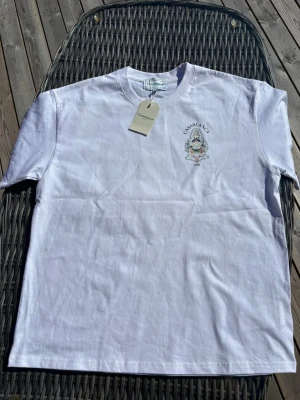 Vit t-shirt från Casablanca - Säljer en fräsch vit t-shirt från Casablanca med tryck på bröstet och rund hals. T-shirten har korta ärmar och är gjord i mjuk bomull. Perfekt för dig som gillar stilrena plagg med en exklusiv touch.