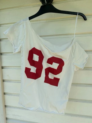 Vit off-shoulder topp med röd 92 - Säljer en vit off-shoulder topp med stort rött tryck av siffran 92 på framsidan. Den är i XXS. Men den passar mig som har S. Så den funkar nog för allt mellan det 🩷