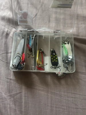 Fiskelåda med drag och beten - Plastlåda med flera olika fiskedrag i metall. Drag i färger som silver, blått, guld, svart och grönt, vissa med prickar och röda detaljer. Perfekt för fiskeentusiasten som vill ha variation på sina beten.