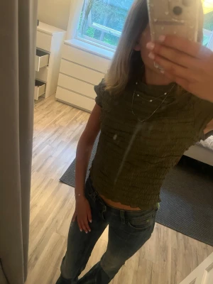 Olivgrön smockad topp från Kappahl - Trendig olivgrön topp från Kappahl i storlek S. Modellen är smockad med korta ärmar och har en croppad passform. Materialet är stretchigt och bekvämt, perfekt till jeans eller kjol. Enkel men snygg detalj med vågig kant nedtill. (Fläcken är på spegeln och inte tröjan, tröjan är aldrig använd bara testad)