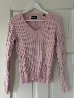 Ljusrosa kabelstickad tröja med v-ringning - Supersöt ljusrosa kabelstickad tröja med v-ringning och långa ärmar. Tröjan har ribbade muddar och ett litet broderat emblem på bröstet. Perfekt att styla med jeans eller kjol för en chill look. Den säljs inte heller längre och är i bra skick.